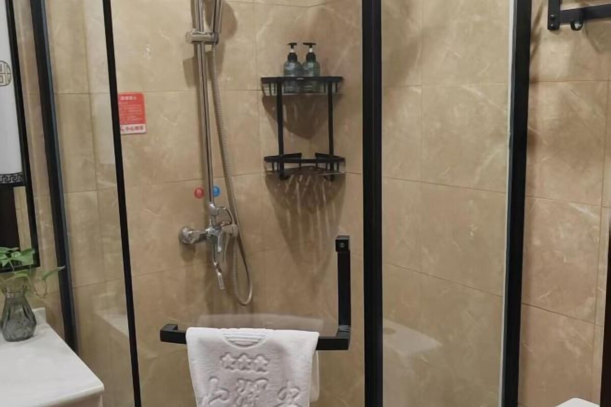 A glass-enclosed shower with a handheld showerhead, a towel rack with a towel, and a shelf with toiletries.