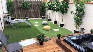A wooden deck with a hot tub, a small table, and chairs. There is a grassy area with a slide and potted plants.