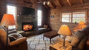 L’intérieur chaleureux d’un chalet avec une cheminée, des murs en bois, un ventilateur de plafond et deux lampes.