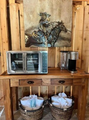 A console table offers a microwave oven and a glass container, accompanied by wicker baskets holding towels and a framed artwork above.