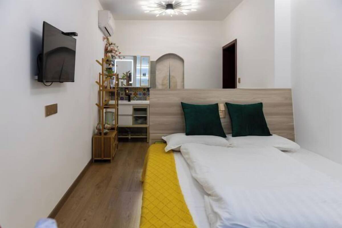 A bedroom with a bed, a TV, a wooden floor, a mirror, and a small plant.