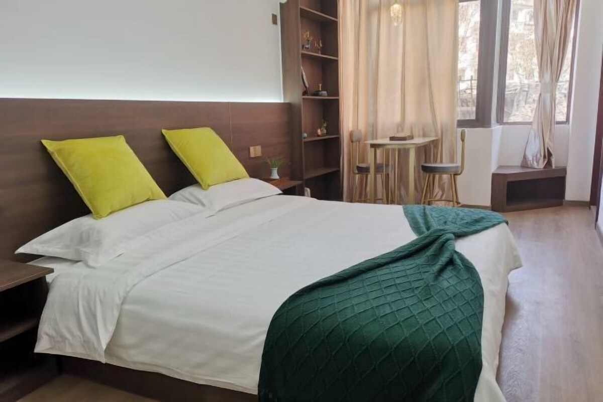A neatly made bed with white bedding and a green throw, a wooden nightstand, a small table with a chair, and a bookshelf.