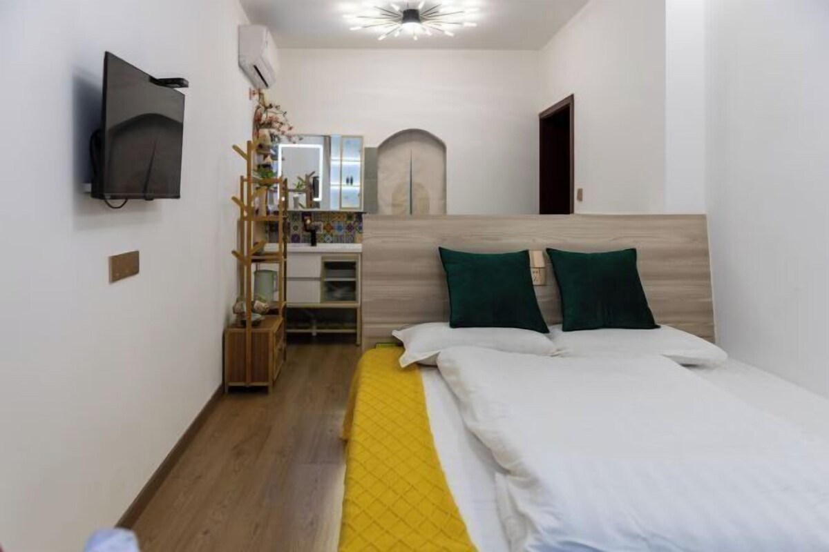 A bedroom with a bed, a TV mounted on the wall, a wooden floor, and a small wooden cabinet.