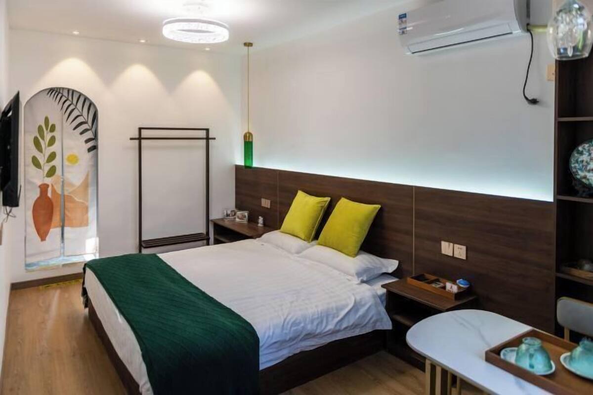 A hotel room with a bed, bedside tables, a desk, and an air conditioning unit.