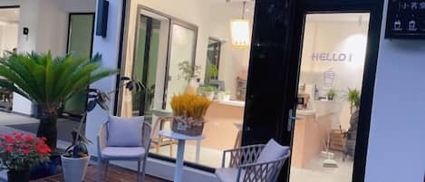 A hotel entrance with a glass door, a 'HELLO!' sign, and outdoor seating with chairs and a table.