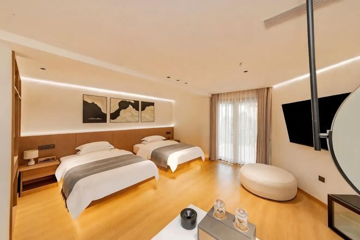 A modern hotel room with two beds, a large mirror, a flat-screen TV, and a bedside table with a lamp.