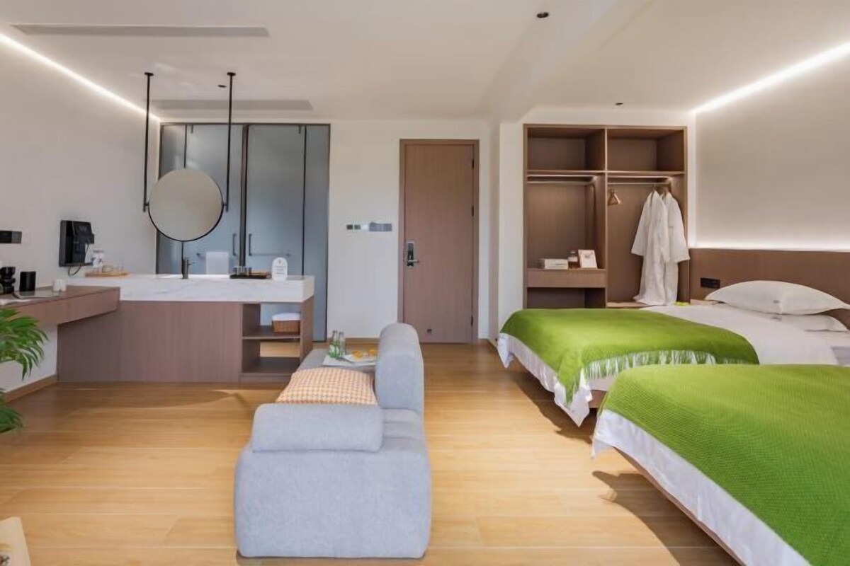 A modern hotel room with a sofa, two beds, a walk-in closet, and a bathroom with a mirror and sink.