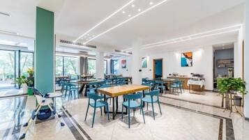 A spacious dining area with multiple tables and chairs, a high ceiling with recessed lighting, and large windows allowing natural light.