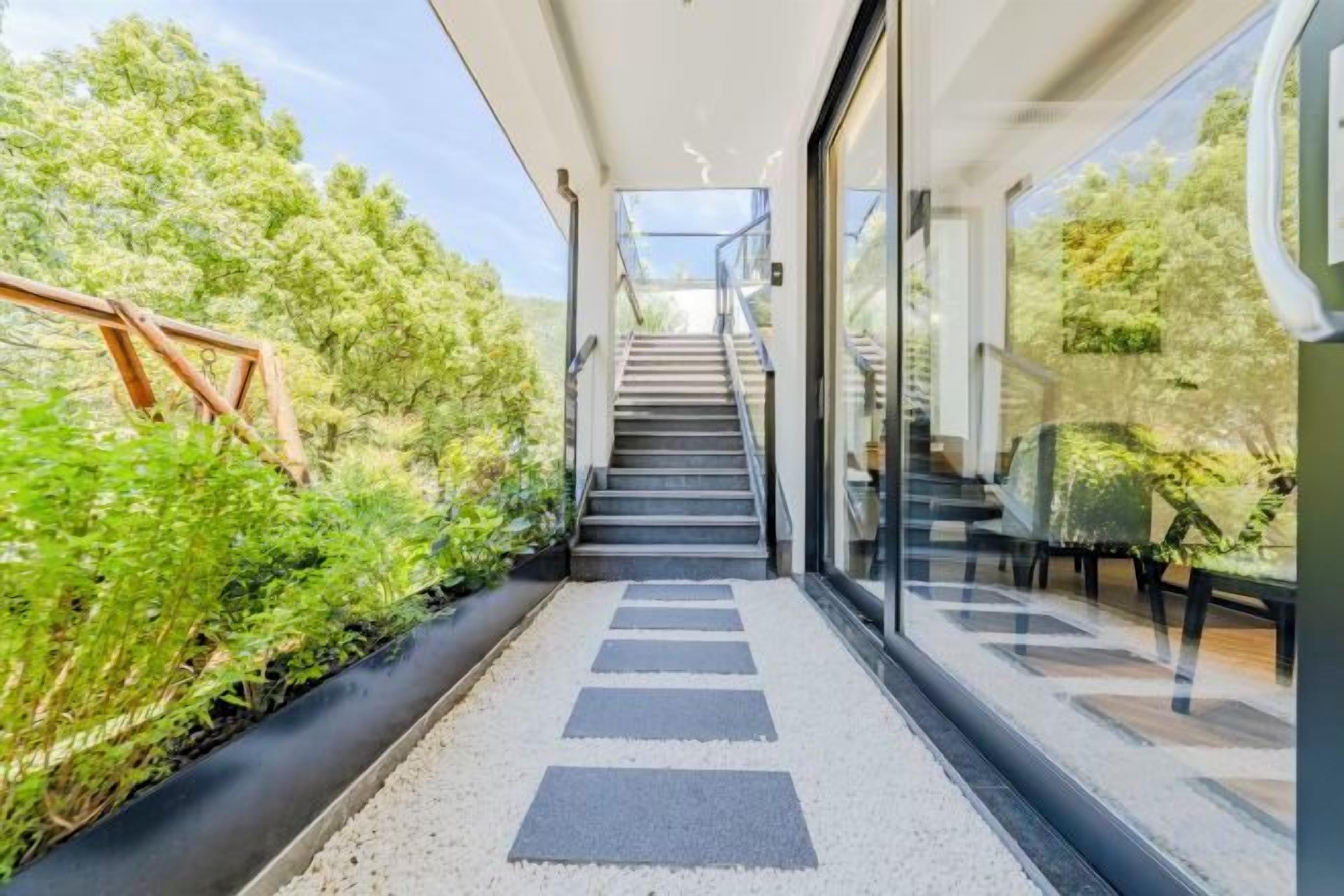 Una escala moderna condueix a una entrada amb porta de vidre, envoltada de vegetació i un cel serè.