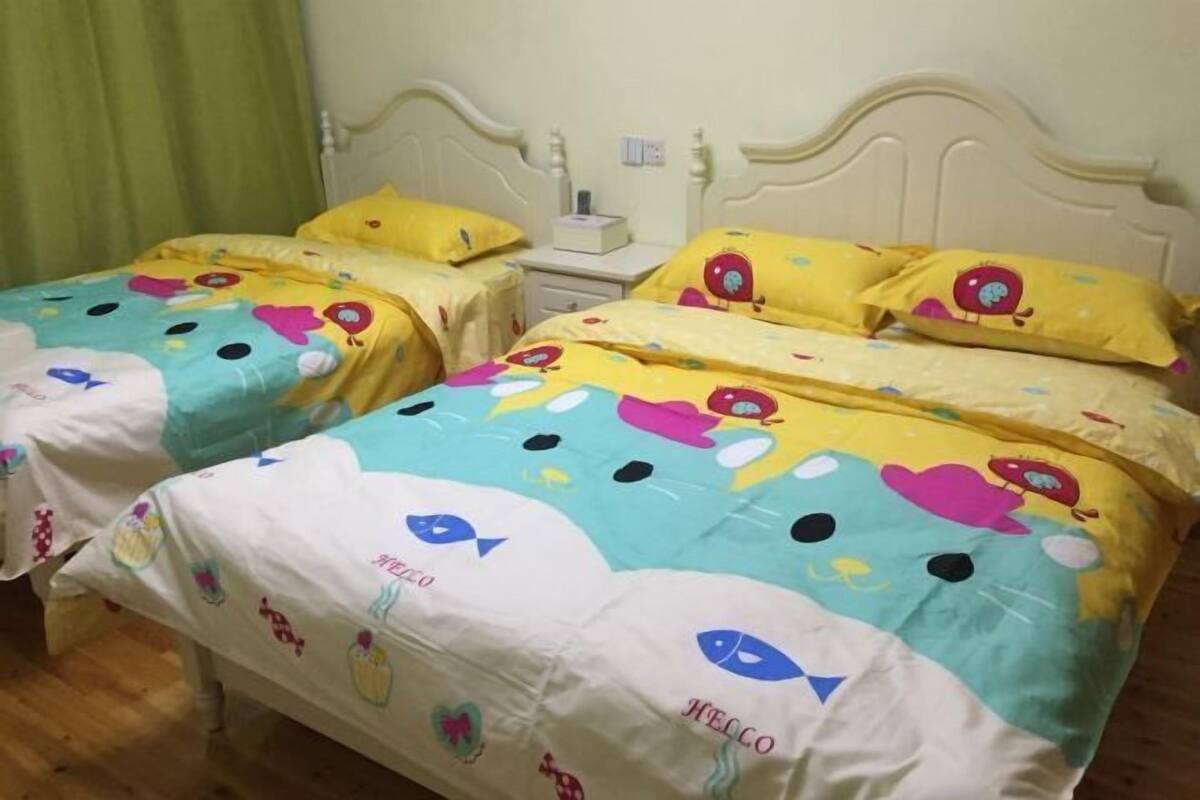 A room with two beds, each with yellow and white bedding featuring cartoon characters. There is a nightstand with a lamp and a small table with a box on it.