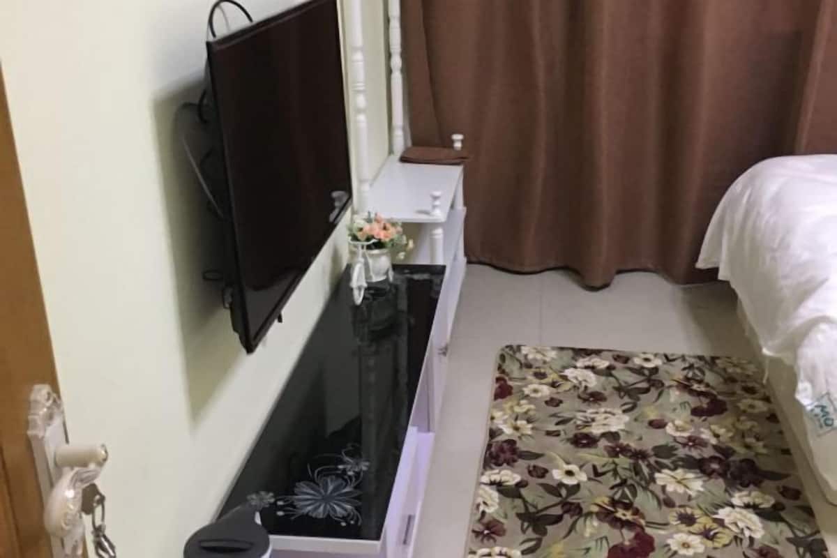 A bedroom with a bed, a TV mounted on the wall, a small bedside table with a vase of flowers, and a floral-patterned rug.