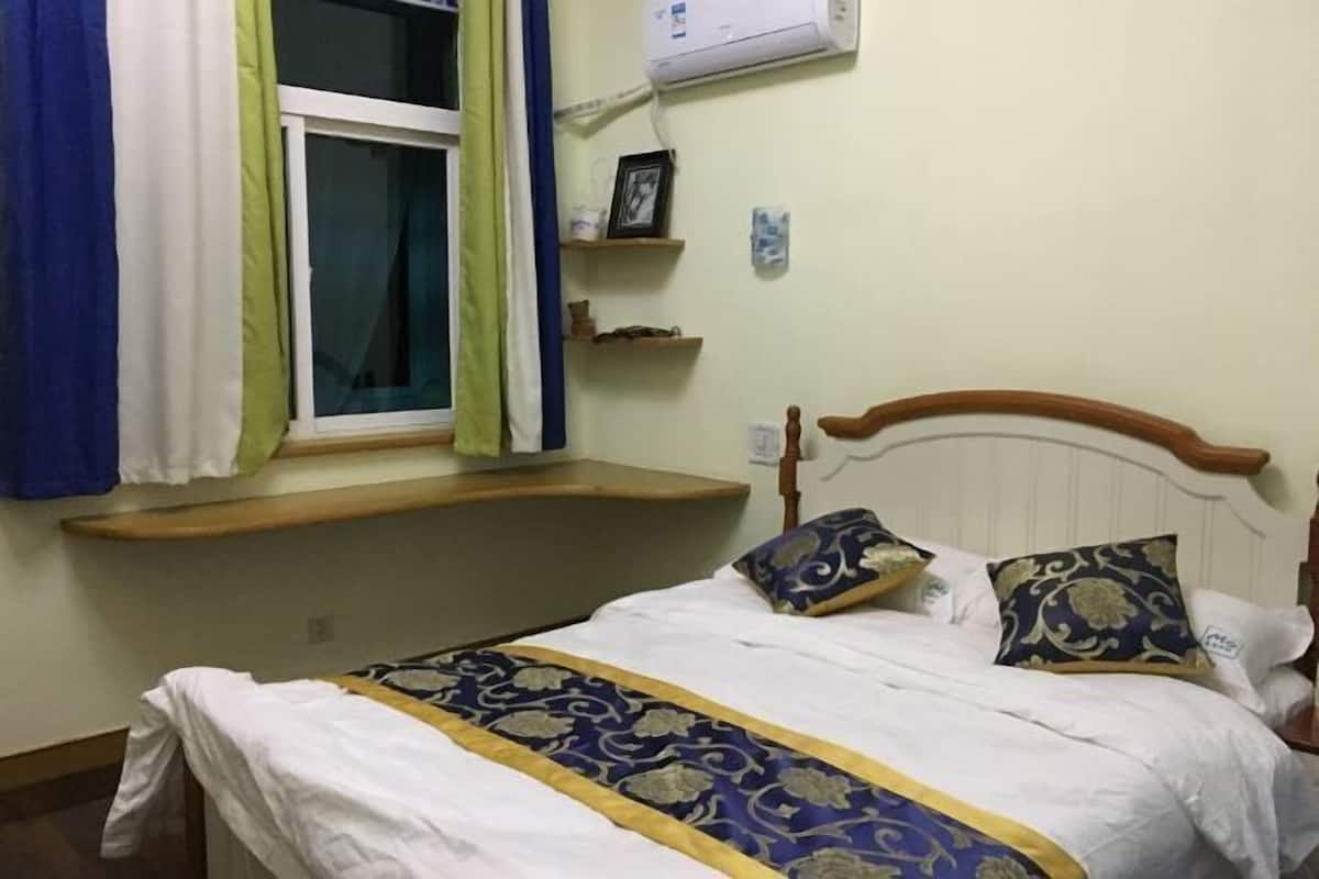 A bed with a patterned bedspread, a window with curtains, a shelf above the bed, and a wall-mounted air conditioner.