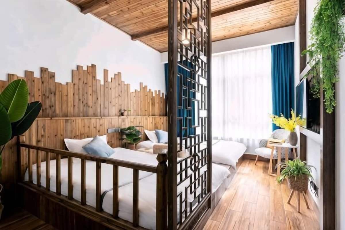 Una habitación con cabecera de madera, cama con ropa blanca, una mesita con una planta y una ventana con cortinas azules.