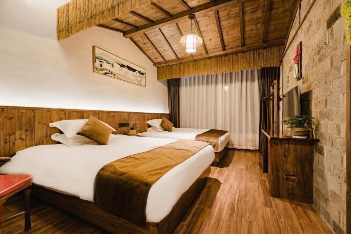 A hotel room with two beds, wooden flooring, a wooden ceiling, a painting on the wall, and a window with curtains.