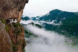 Un petit bâtiment perché au bord d’une falaise, offrant une vue sur un paysage montagneux et sur les nuages en contrebas.