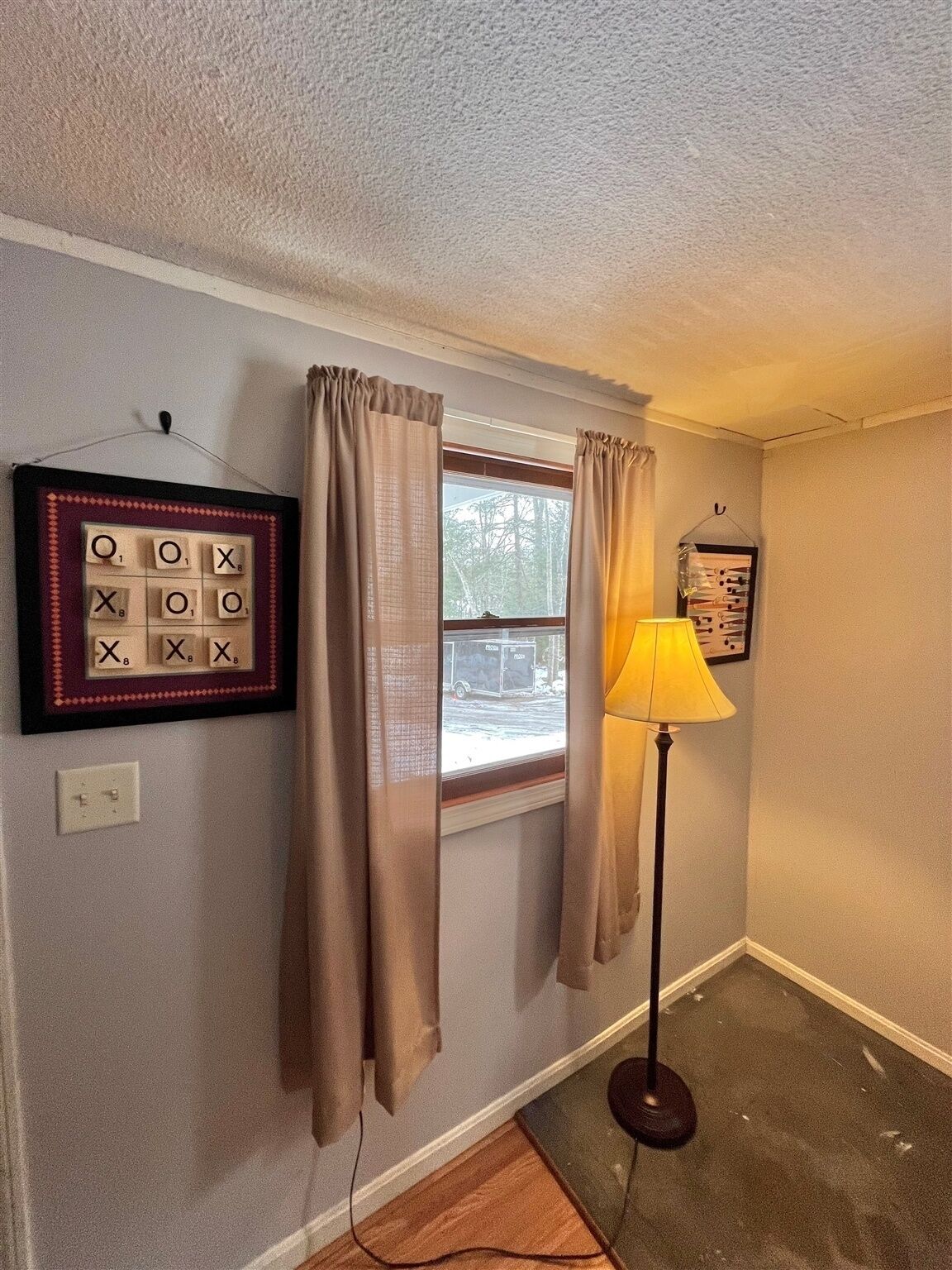A room with a window, curtains, a framed Scrabble game, a lamp, and a power outlet.