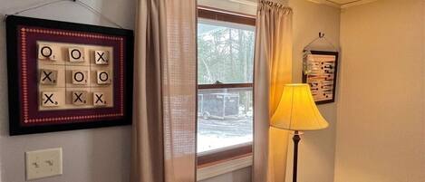 A room with a window, curtains, a framed Scrabble game, a lamp, and a power outlet.