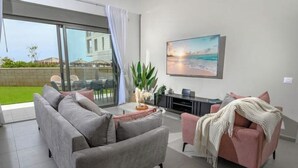 Een moderne woonkamer met een grijze bank, een roze fauteuil en een salontafel. Er is een groot raam met uitzicht op een tuin en een gebouw. Op de muur hangt een schilderij van een strand.