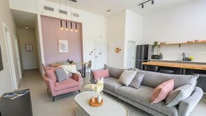 Een moderne woonkamer met een grijze bank, een roze fauteuil en een witte salontafel. Er is een keukenhoek met een magnetron en een houten aanrecht.