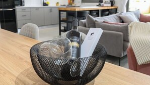 Een keuken met een eettafel, een zwarte netmand met een fles en glazen, en een bank op de achtergrond.