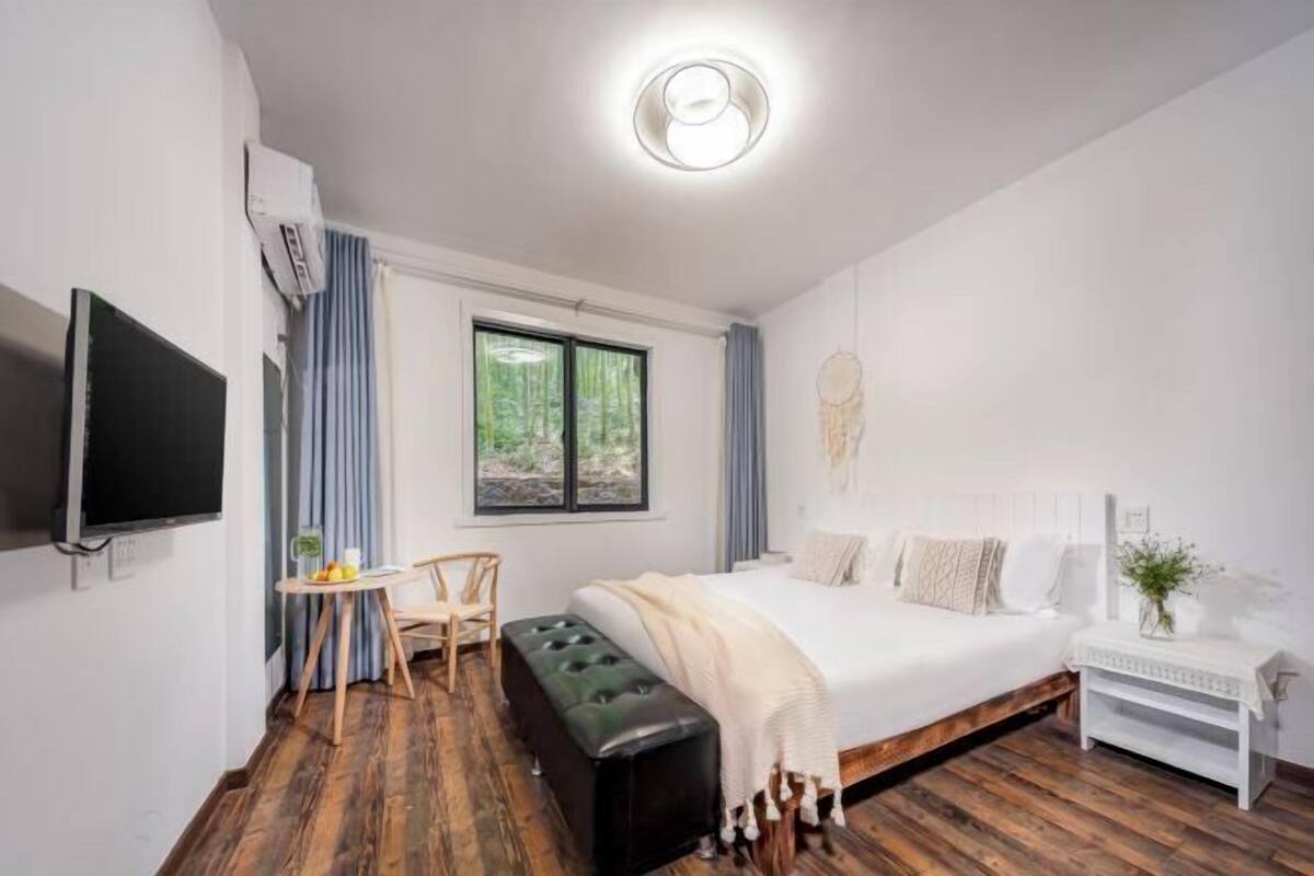 Un dormitori modern amb un llit gran, una televisió, una taula petita amb una cadira i una finestra amb cortines.