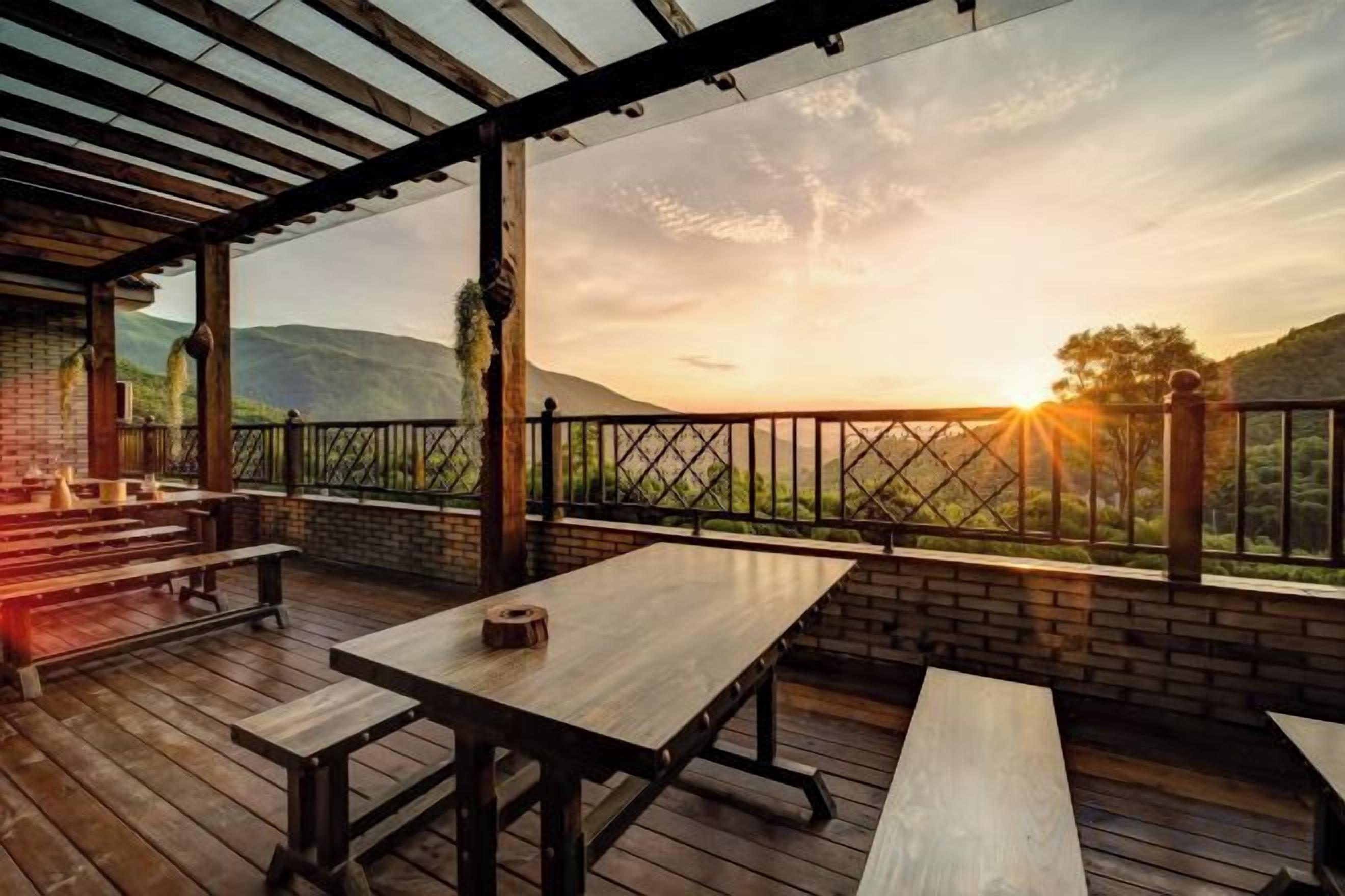 An outdoor patio with wooden tables and benches, a brick wall, and a view of the sunset over a hilly landscape.