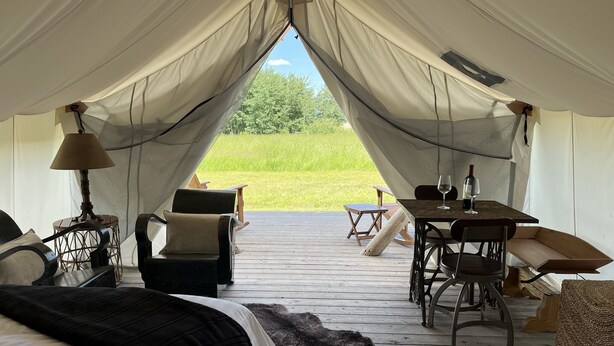 Ett tältliknande boende med en säng, stolar, ett bord och utsikt över en gräsklädd äng.