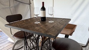 Ett träbord med en vinflaska och två glas, placerat på ett trädäck under ett tält.
