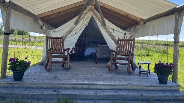 A wooden deck with two rocking chairs and a small table, under a large tent structure. - Heritage Ranch 'Rainbow' Glamping Tent (12) (Bozeman)
