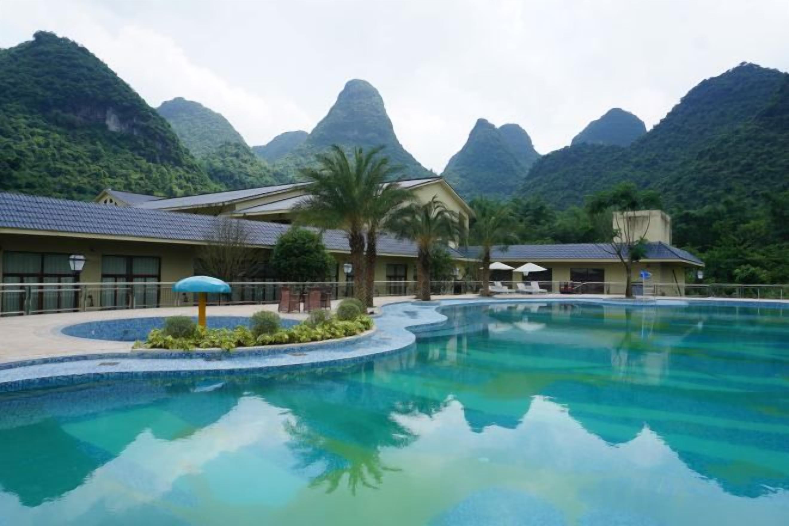 A resort with a swimming pool, palm trees, and mountainous backdrop.