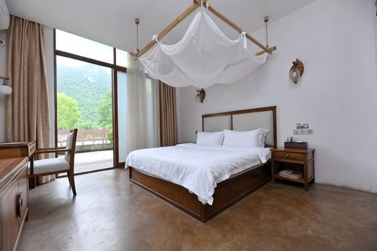 A bedroom with a large bed, a nightstand, a chair, and a window with a view of greenery.