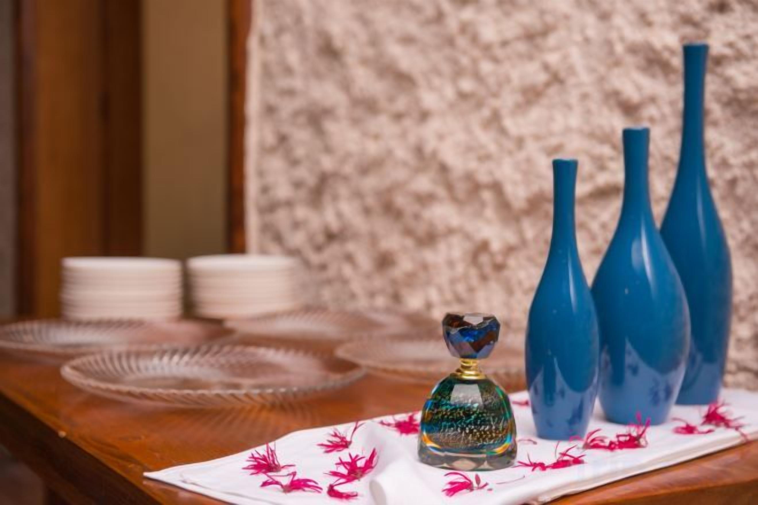 Een tafel met een decoratieve glazen fles en drie blauwe vazen, voor een rustieke wand.