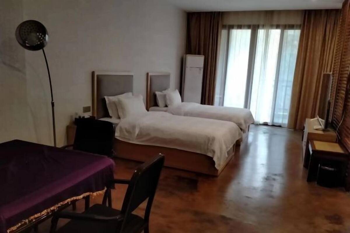 A hotel room with two beds, a desk, a chair, a lamp, and a view of the outdoors through large windows.
