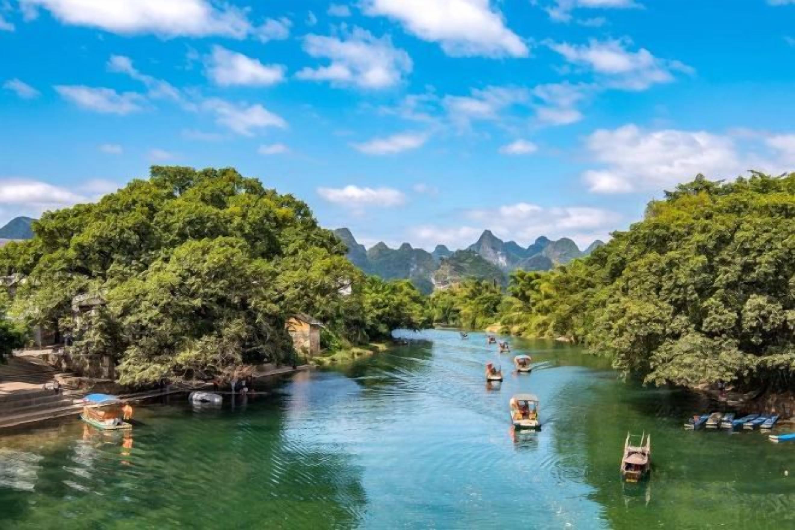 A river with boats, surrounded by lush greenery and mountains in the background.
