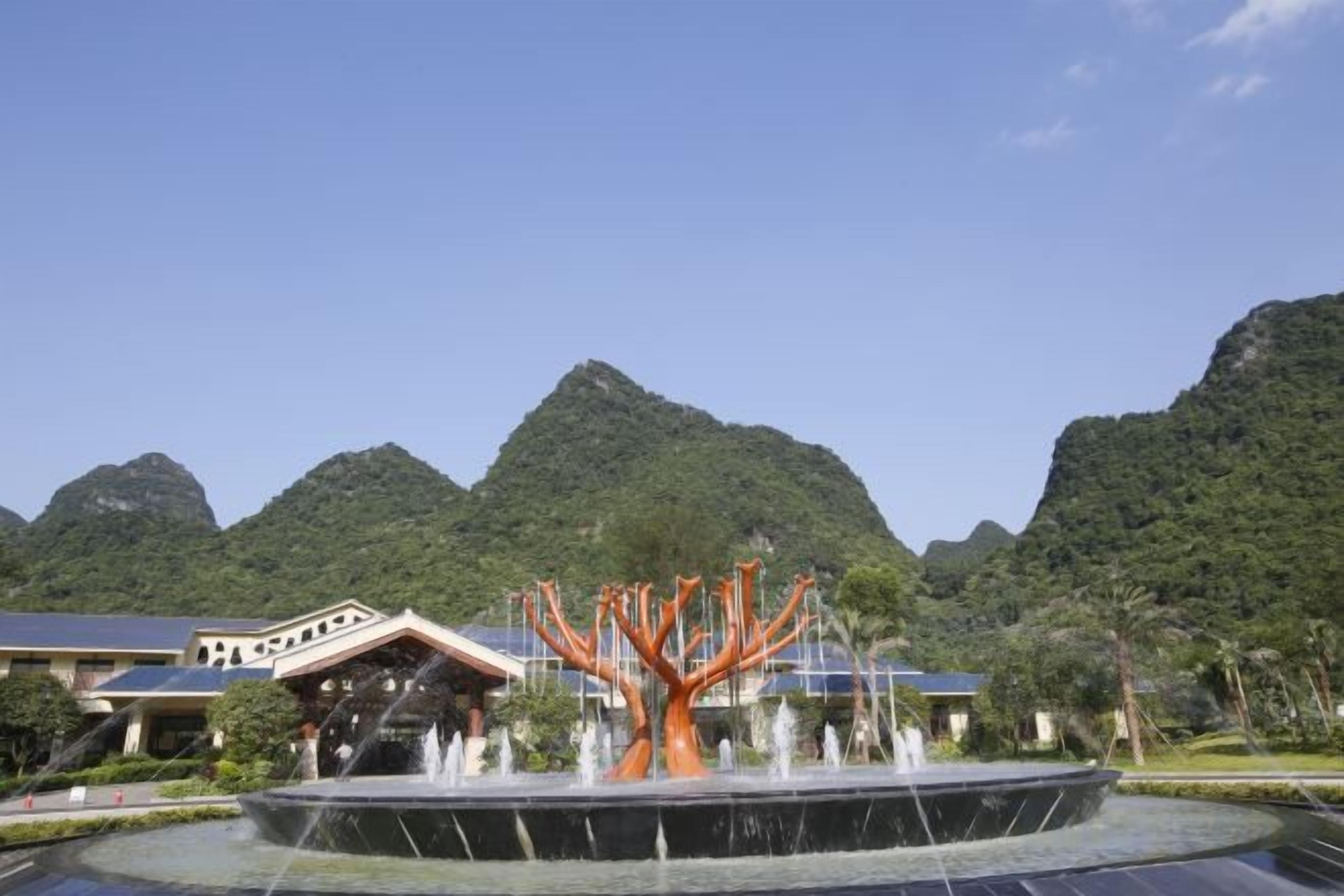 A fountain with a tree-like structure in the center, surrounded by a pool, with mountains in the background.