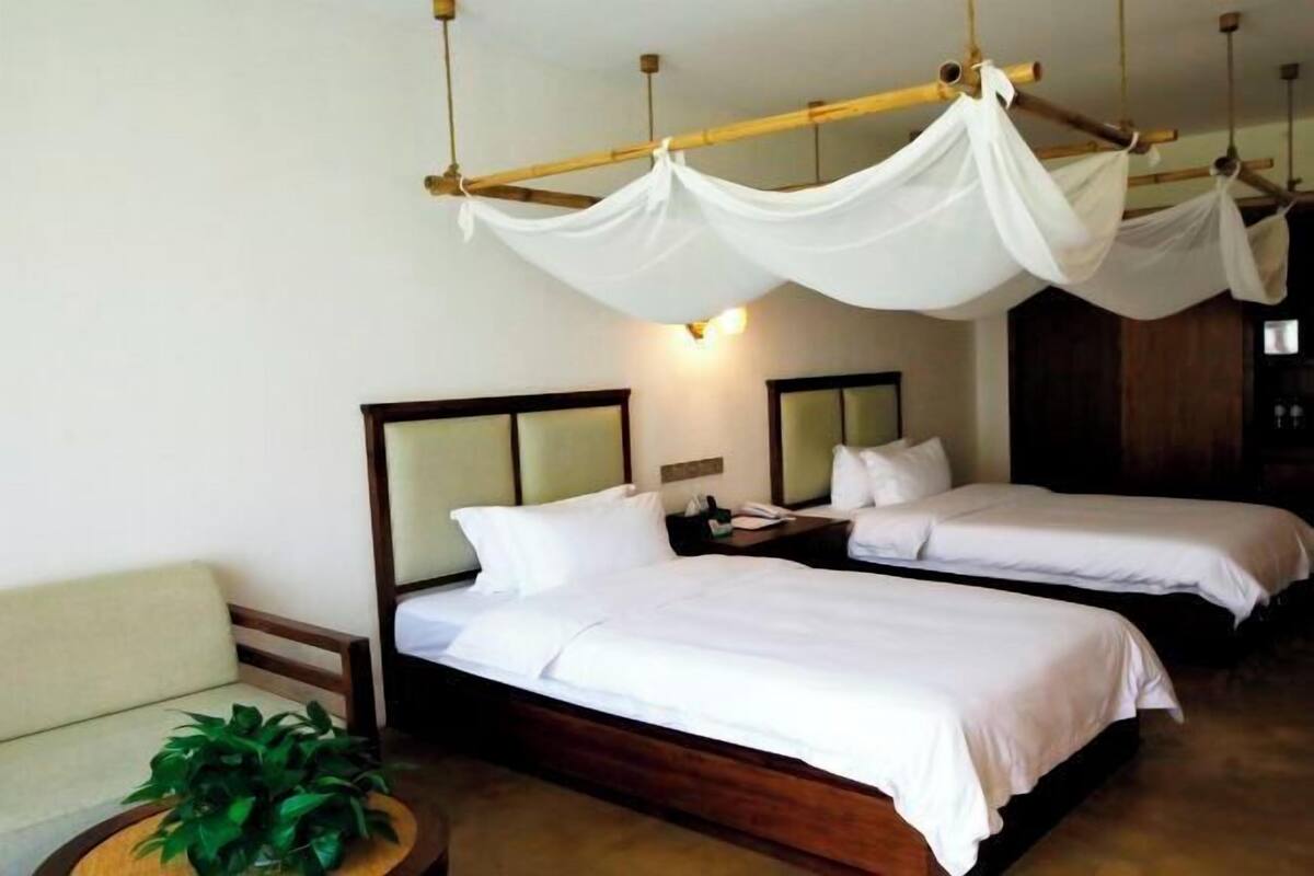 A hotel room with two beds, a wooden headboard, a bedside table with a lamp, and a small round table with a plant.