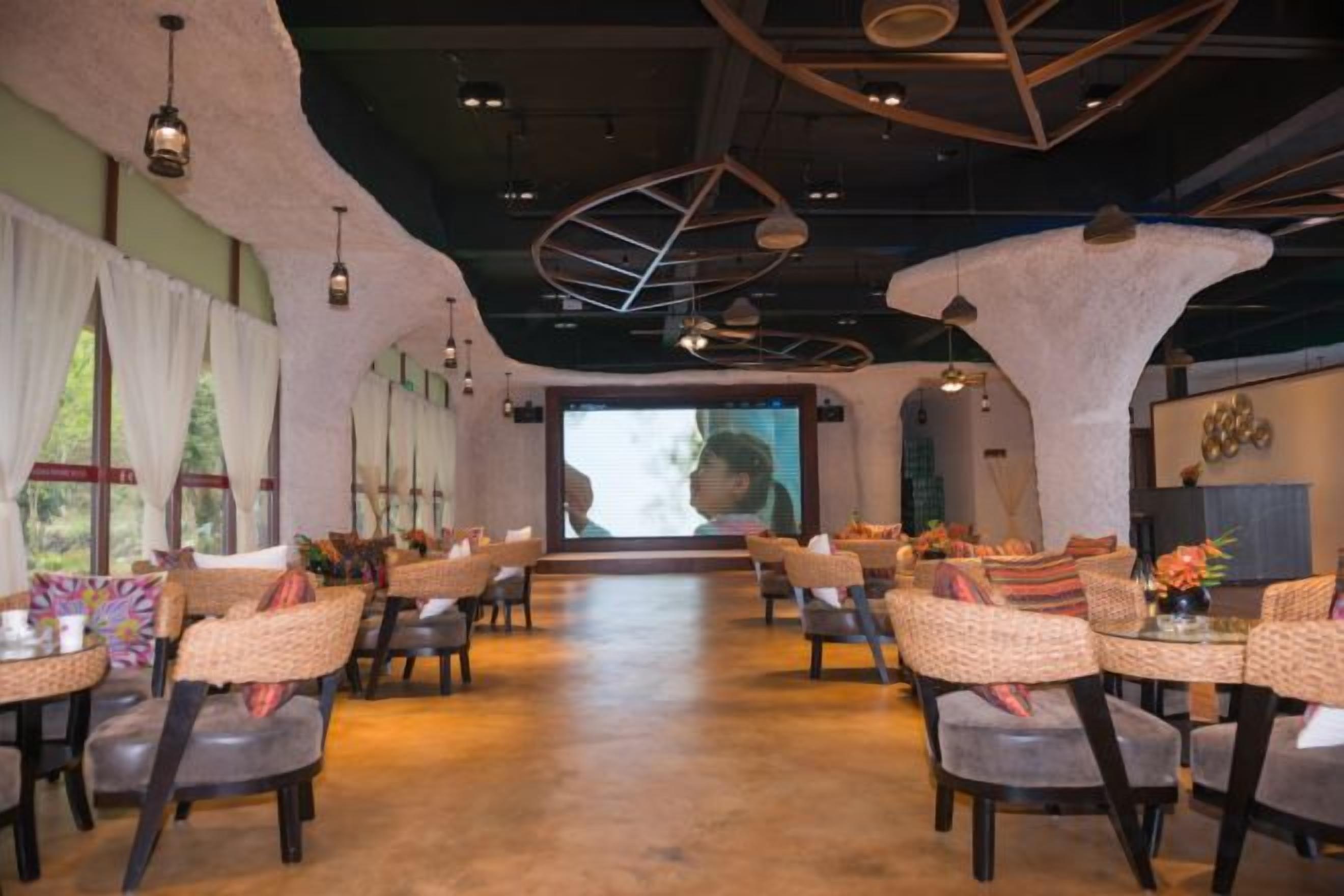 A spacious dining area with wicker chairs and tables, a large screen at the front, and a modern ceiling design.