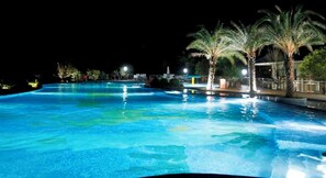 Piscina noturna com água cristalina, rodeada por palmeiras e iluminada por iluminação ambiente.