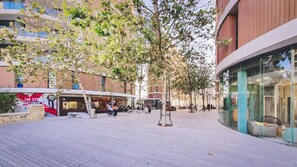 An urban pedestrian area with a wooden boardwalk, trees, and modern buildings with shops and cafes.