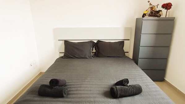 A bed with a grey bedspread, two pillows, and two rolled towels on the bed.