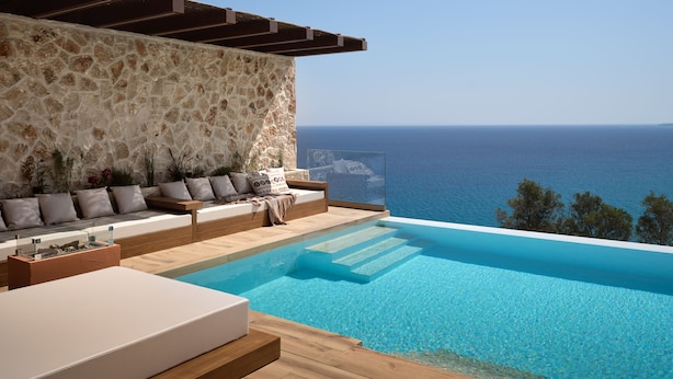 Un área de piscina moderna al aire libre con muros de piedra y una terraza de madera, con vistas al mar azul y cristalino.
