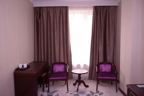 Ein Hotelzimmer mit Esstisch, zwei violetten Stühlen, einem kleinen runden Tisch und Blick durch ein Fenster mit Vorhängen.