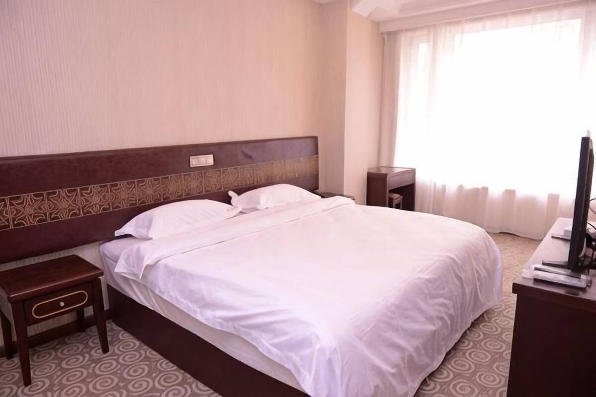 Egy szállodai szoba, amelyben egy nagy ágy, egy éjjeliszemélyzet, egy televízió és egy függönnyel ellátott ablak található.