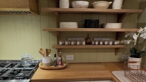 Una cocina con estufa a gas, horno, armarios blancos, encimera de madera y estantes con vajilla y frascos.