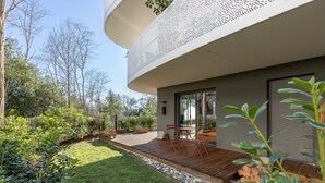 Una casa moderna con terraza de madera, puerta de cristal y jardín con diversas plantas y una mesa de madera.