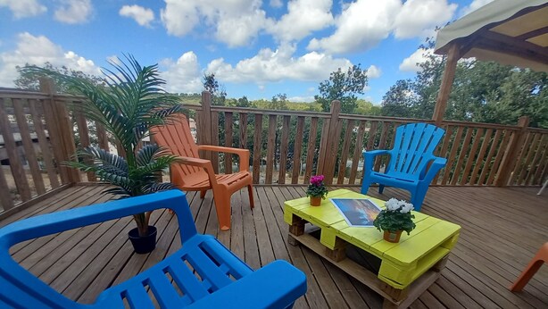 A wooden deck with outdoor furniture including blue and orange chairs, a yellow table, and potted plants. - MOBIL HOME 40 m², 4 persons in Provence Verte. (Rocbaron)