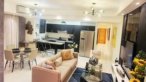 Una sala moderna con sofá, mesa de centro y comedor. Se observa una cocina al fondo con armarios y electrodomésticos.