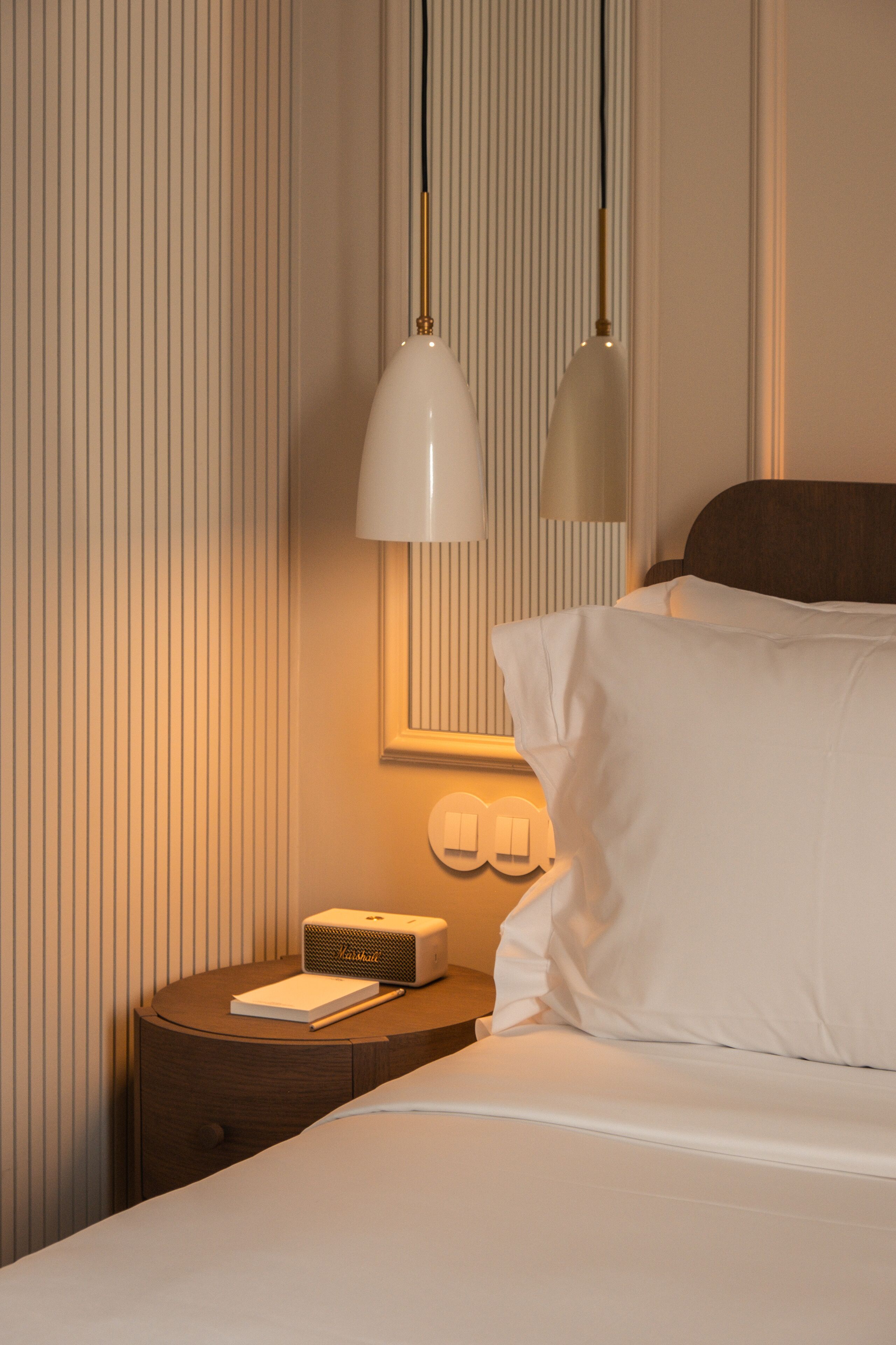A neatly made bed with white linens, a bedside lamp, a small wooden nightstand with a book and a device, and a wall with vertical lines.