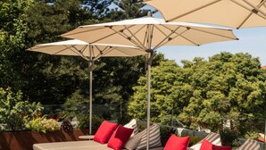 A deck with wooden flooring, beige cushioned seating with red and striped pillows, and beige umbrellas providing shade.