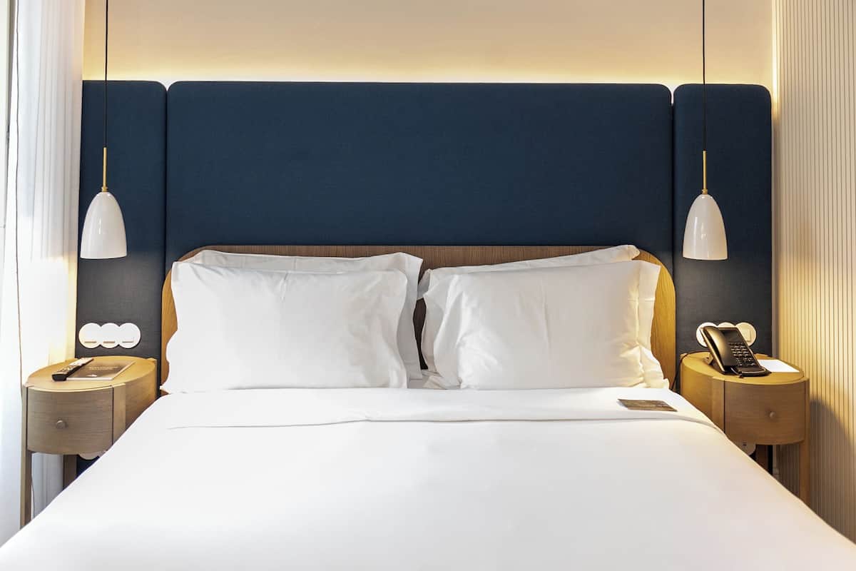 Quarto de hotel com uma cama grande, duas mesas de cabeceira, telefone ao lado da cama e luminária fixada na parede.
